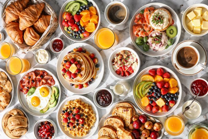 Overhead view of a breakfast table with cereal, granola, orange juice, yogurt, and toast — common breakfast foods that contain hidden added sugar